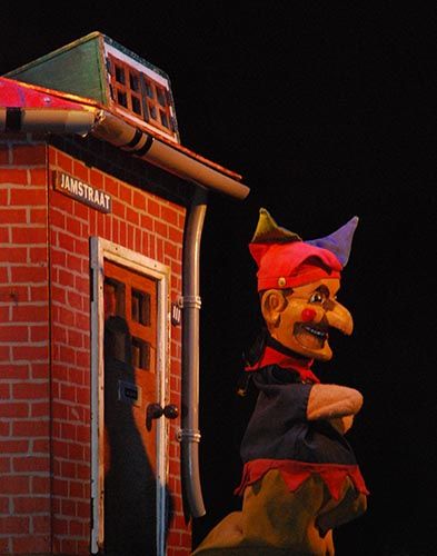 Poppenkastpop Jan Klaassen voor een huisje met een donkere achtergrond.