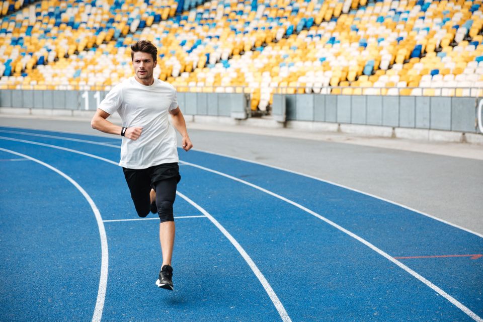 Atletiekbaan met hardloper tijdens een training, met een sporter in hardloopkleding op een blauwe renbaan en kleurrijke stadiontribunes op de achtergrond.