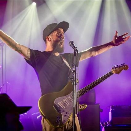 Muziekoptreden met gitarist en zanger op verlicht podium tijdens live concert, artiest met elektrische gitaar en microfoon, sfeerbeeld van optreden met podiumverlichting en publiekservaring.