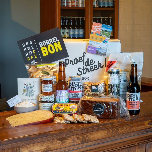 Inhoud van de Proef de Streek Kerstbox. Je ziet onder andere flesjes bier van Pronck, Katwijkse Knip koekjes, ontbijtkoek, een stuk Leidse Kaas, borrelnootjes, blikje sardientjes, een cadeaubon en een kartonnen kerspakketdoos van Proef de Streek.