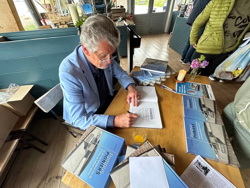 Een man zit achter een tafel met een stapel boeken en zet zijn handtekening in de boeken.