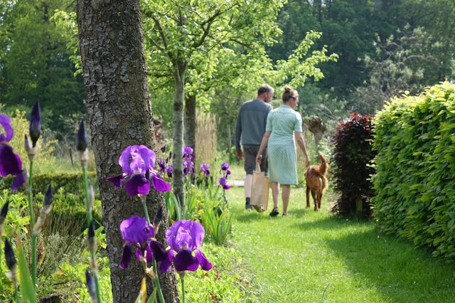 Twee mensen wandelen met een hond door een tuin. Op de voorgrond zie je paarse bloemen.