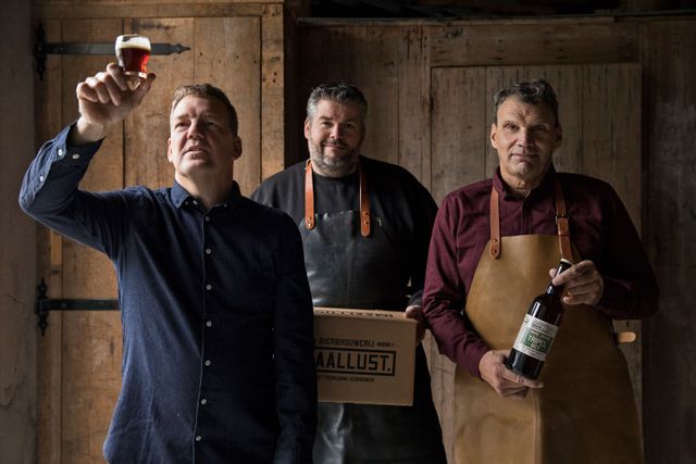 Drie mannen van brouwerij Maallust. De een houdt een biertje tegen het licht, de middelste heeft een doos vast met het logo van Maallust erop en de derde heeft een flesje bier vast.
