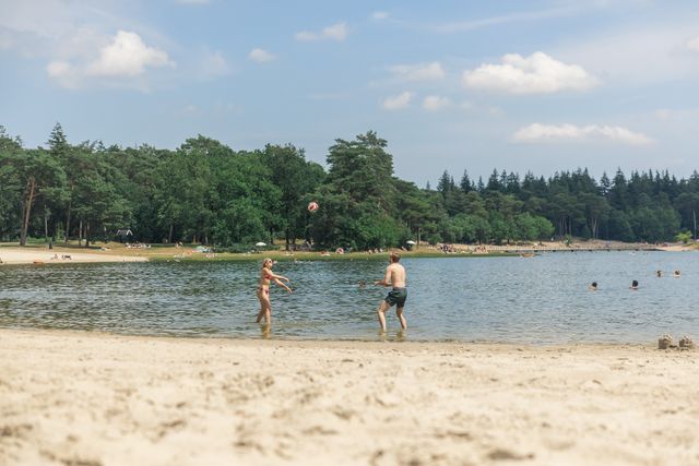 Twee kinderen gooien over met een bal op het strand van het Henschotermeer. Er is water en bos en de zon schijnt.