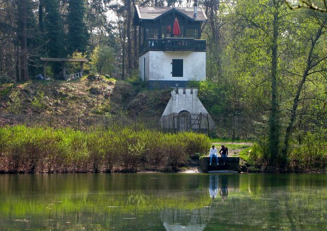 Historisch theehuis aan de vijver op Landgoed Heidestein bij Zeist, omringd door bos en groen. Twee bezoekers zitten aan de waterkant met uitzicht op het karakteristieke gebouw.