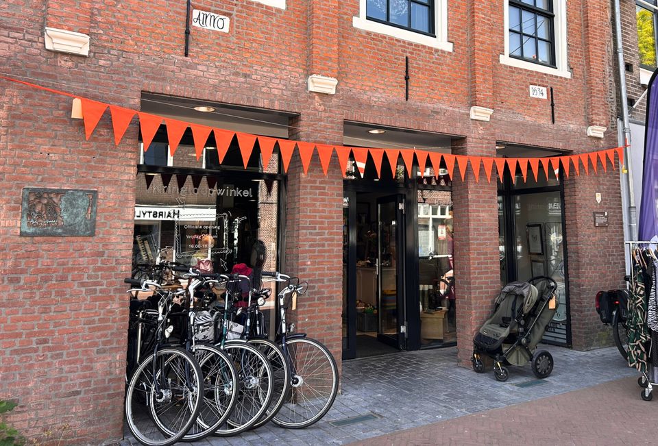 Voorgevel van kringloopwinkel De Terugwinning in het centrum van Woerden, met oranje vlaggetjes, fietsen voor de deur en kledingrekken buiten op straat.