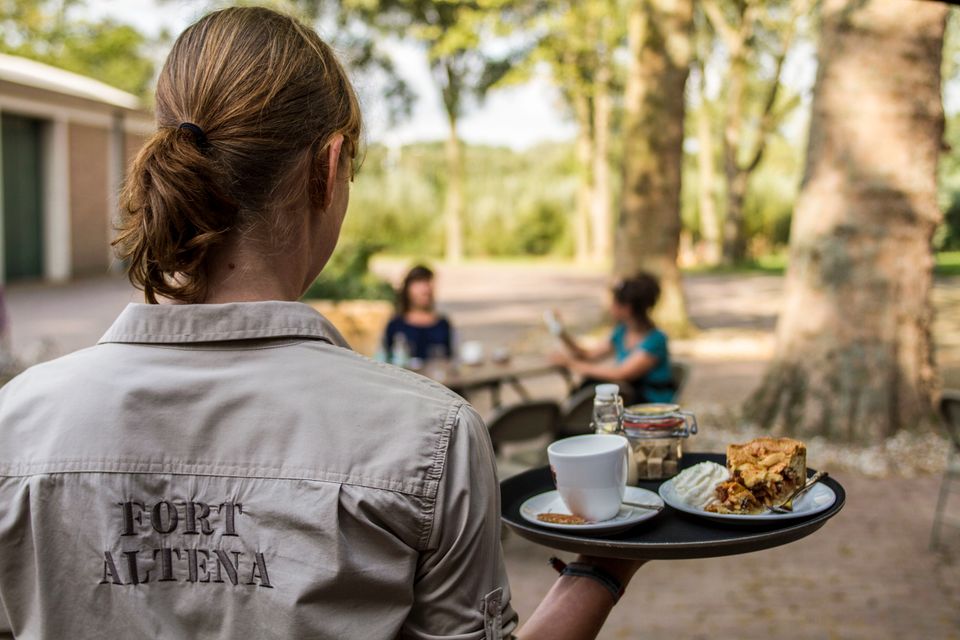 Een vrouw met een bruin overhemd met op de rug de woorden 'Fort Altena' bedient op een terras. Op haar dienblad staat thee en een punt appeltaart.