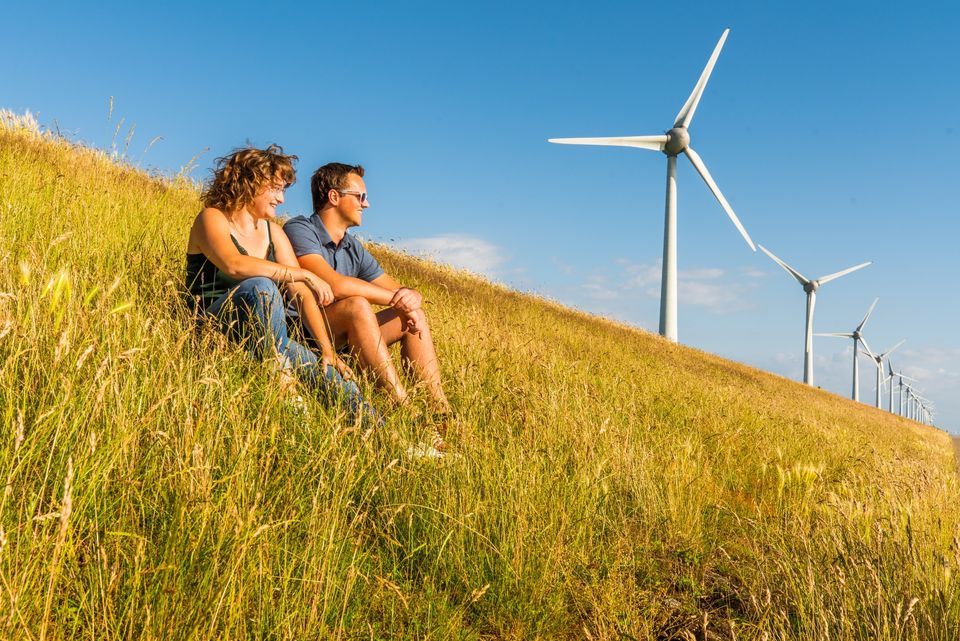 Een stel zit naast elkaar op een dijk en kijkt voor zich uit met op de achtergrond windmolens uit de Noordoostpolder.