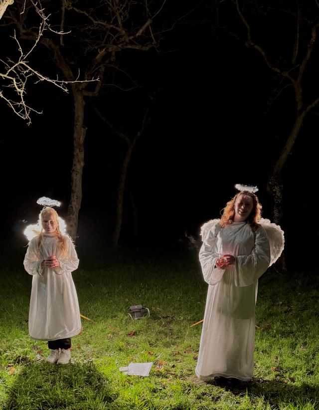 Twee verklede engelen staan in een nachtelijk bos tijdens een kaarslichtwandeling. Ze dragen witte gewaden, vleugels en halo’s, terwijl zachte verlichting hen omringt en een serene, magische sfeer creëert.