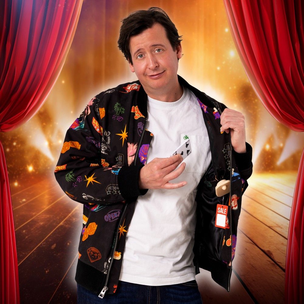 Steven Kazàn in een staat op een podium tussen 2 open gordijnen. Hij heeft een kleurrijk jasje aan met een paar speelkaarten in zijn handen.