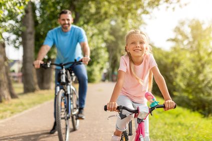 Meisje fietst voorop over een zonnig pad door het groen, terwijl een man op een fiets achter haar volgt; samen genieten ze van een ontspannen fietstocht in de natuur.