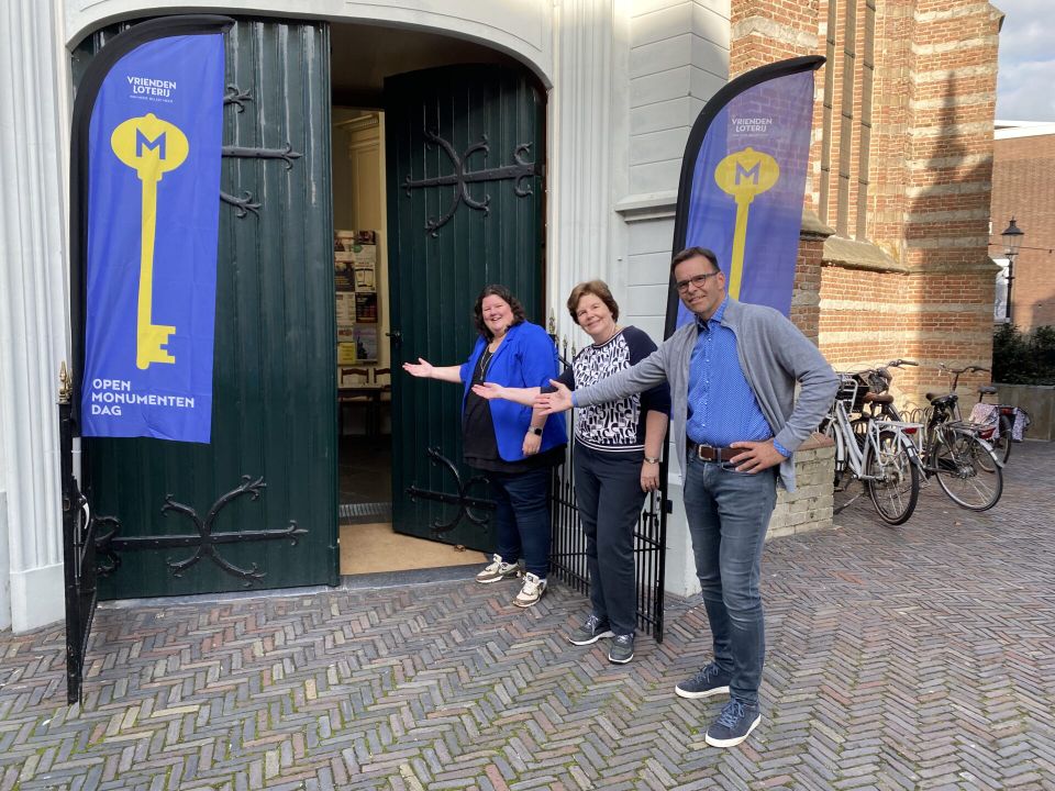Drie vrijwilligers verwelkomen bezoekers bij de ingang van een monumentale kerk tijdens Open Monumentendag in Woerden, met vlaggen aan weerszijden van de deur.