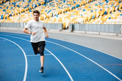Atletiekbaan met hardloper tijdens een training, met een sporter in hardloopkleding op een blauwe renbaan en kleurrijke stadiontribunes op de achtergrond.