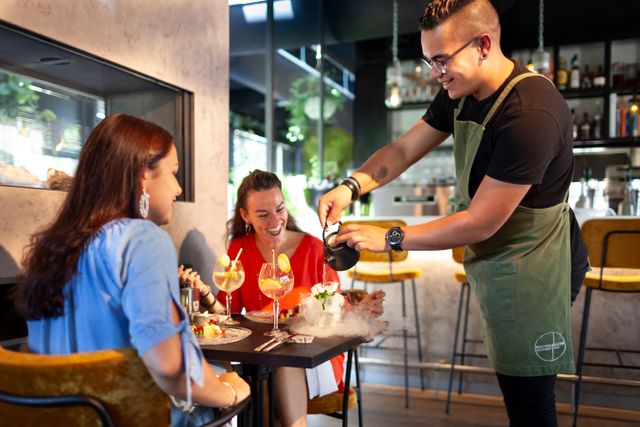 Twee vriendinnen genieten bij Bistronoom in Woerden terwijl een gastvrije ober een creatief gerecht met rookeffect serveert; een culinaire beleving in een moderne, sfeervolle setting in het Groene Hart.