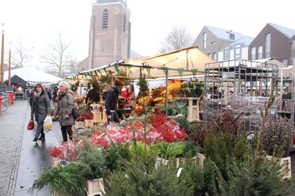 Een sfeervol beeld van de gezellige wintermarkt in Woerden, waar bezoekers tussen kleurrijke bloemen, kerstgroen en winterse decoraties slenteren. De warme lichtjes en het rumoer van kraampjes geven het plein een levendige, feestelijke uitstraling, terwijl de imposante kerk op de achtergrond waakt over het winterse stadsleven.