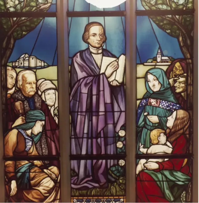 Glas-in-loodraam in Petruskerk Woerden met Jan de Bakker die predikt voor toehoorders, omgeven door bomen, kerken en symboliek uit zijn tijd.