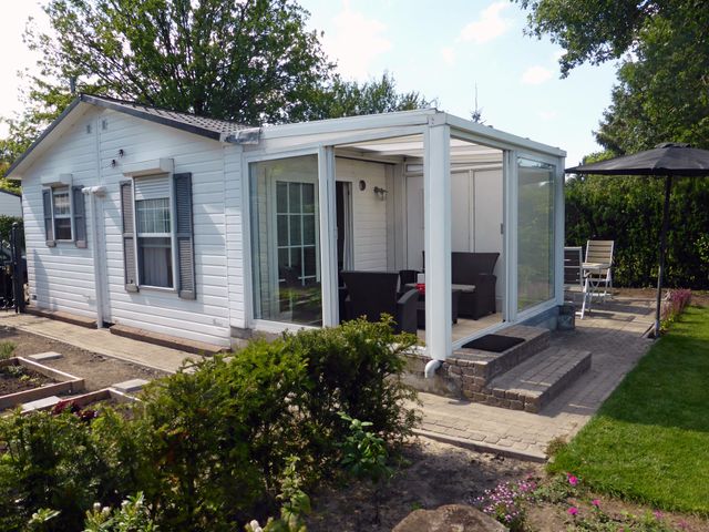 Zicht op de achterkant van een wit houten chalet met grijze luikjes en aan de rechterkant een glazen serre. Terras met een parasol en een zitje en op de voorgrond een moestuintje.