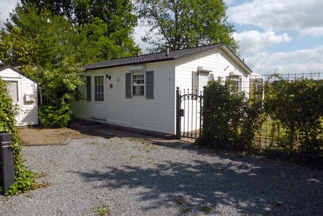 Een witte houten chalet met grijze luikjes aan een grijs grindpad. Boomrijke omgeving en een smeedijzeren hekje geeft toegang tot een tuin.