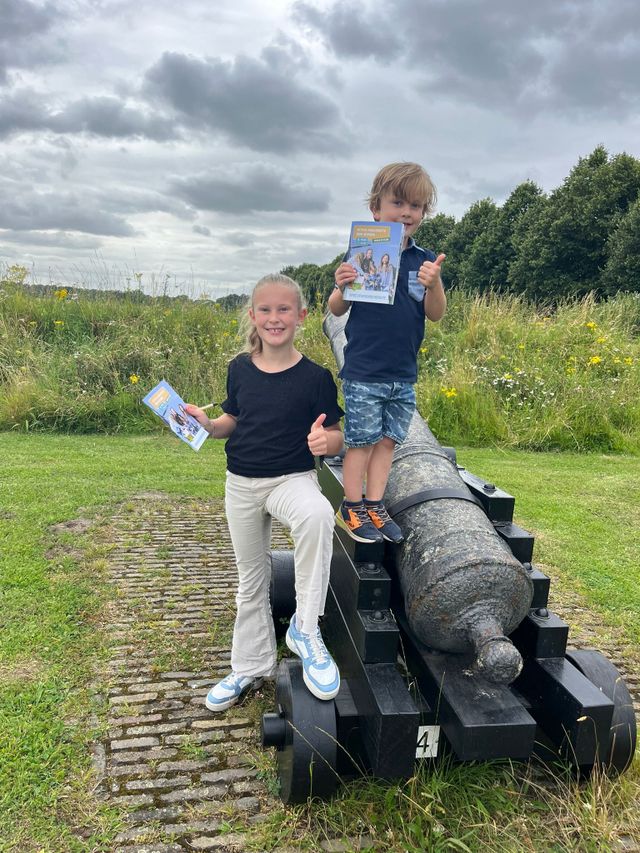 Zwei Kinder stehen mit dem Sprichwortweg-Aufgabeheft in der Hand in der Nähe einer Kanone in der Festung Heusden.