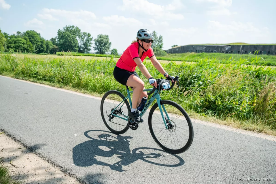 Een vrouw in een rode wielrenoutfit op een wielrenfiets. Op de achtergrond een betonnen fort en groene omgeving.
