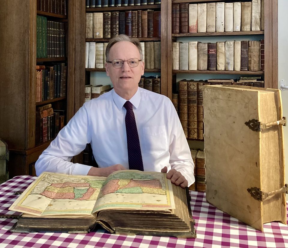 Arie Molendijk zit aan tafel op een kantoor omgeven met een gevulde boekenkast op de achtergrond en op tafel een dicht en opengeslagen oud boek die hij aan het taxeren is.