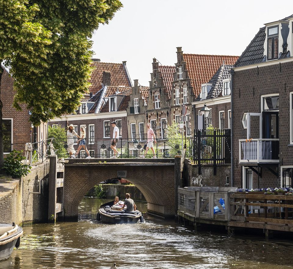 Historische binnenstad Oudewater, Groene Hart. Wandelaars steken een brug over terwijl een sloep onder de brug door vaart over de Hollandse IJssel, met monumentale gevels en grachtenpanden langs het water.