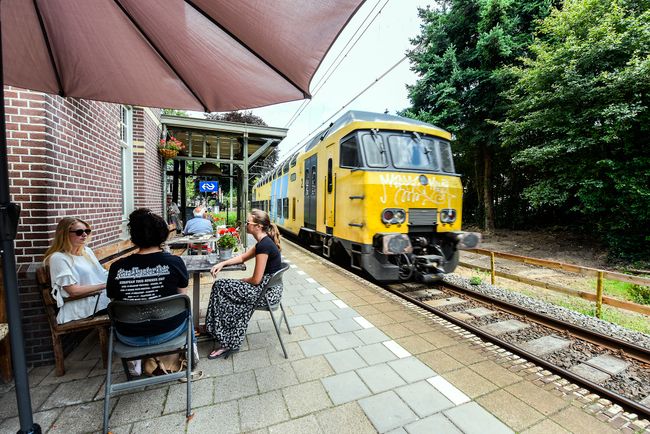 Bezoekers zitten op een terras naast het spoor in Baarn terwijl een trein voorbijrijdt. De foto combineert horeca, spoorhistorie en de bijzondere ligging van het terras.