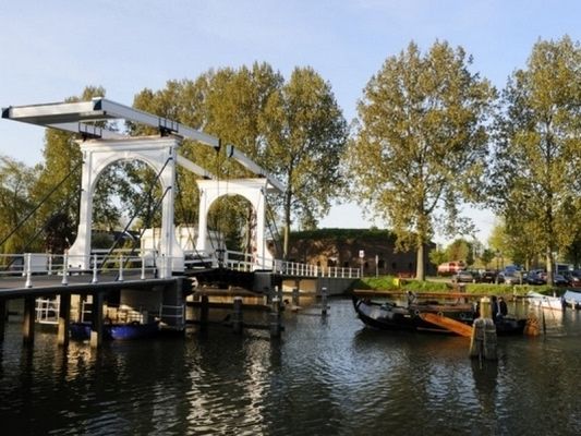Een witte brug over een waterpartij met in de achtergrond een bakstenen fort, omgeven door bomen.