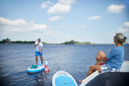 En man staat op een SUP-board op het water en lacht naar een vrouw die op een boot zit.