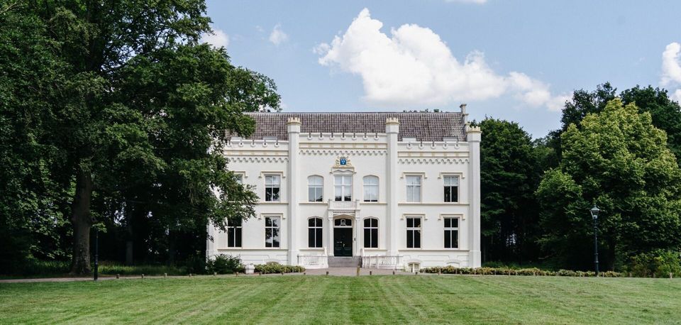 Vooraanzicht van Huis Scherpenzeel, een wit historisch landhuis op een groen landgoed in Scherpenzeel. Het gebouw wordt omringd door oude bomen en een ruim gazon.