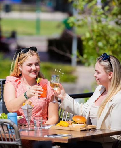 Twee jonge vrouwen proosten met een cocktail en een glas witte wijn op het zonovergoten terras van Proeflokaal Bregje in Woerden, met burgers en friet op tafel in de zon.