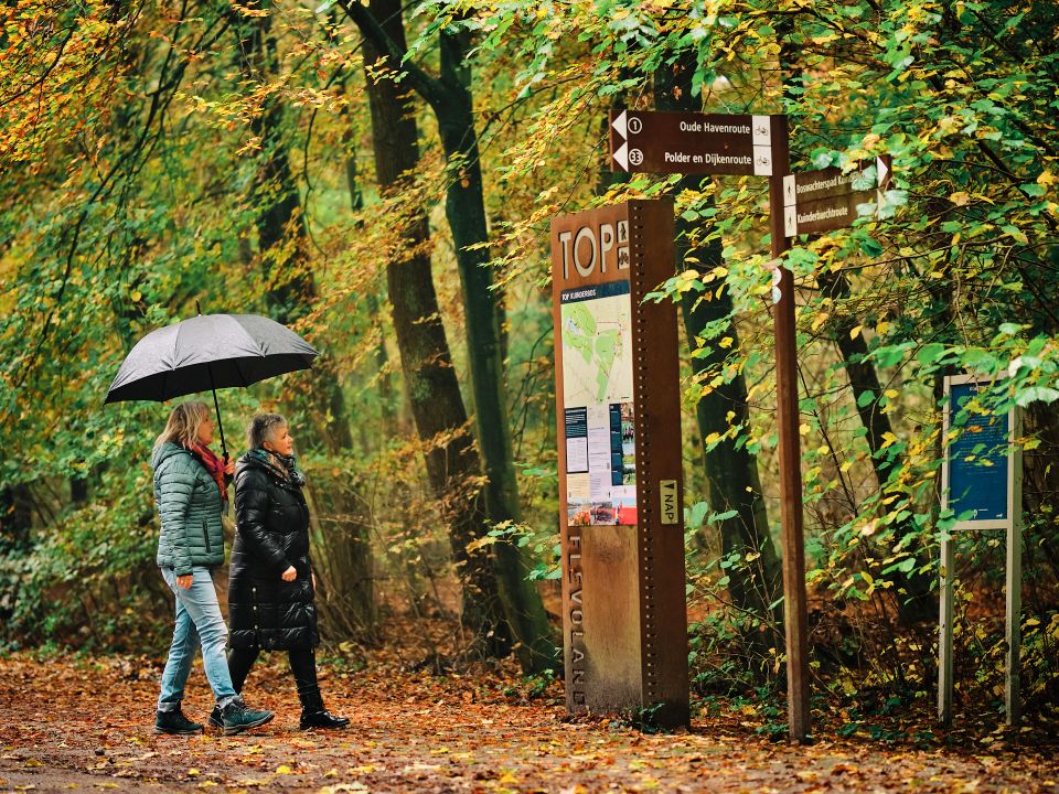 Twee vrouwen staan in een herfstig Kuinderbos bij de TOP een wandelroute uit te zoeken