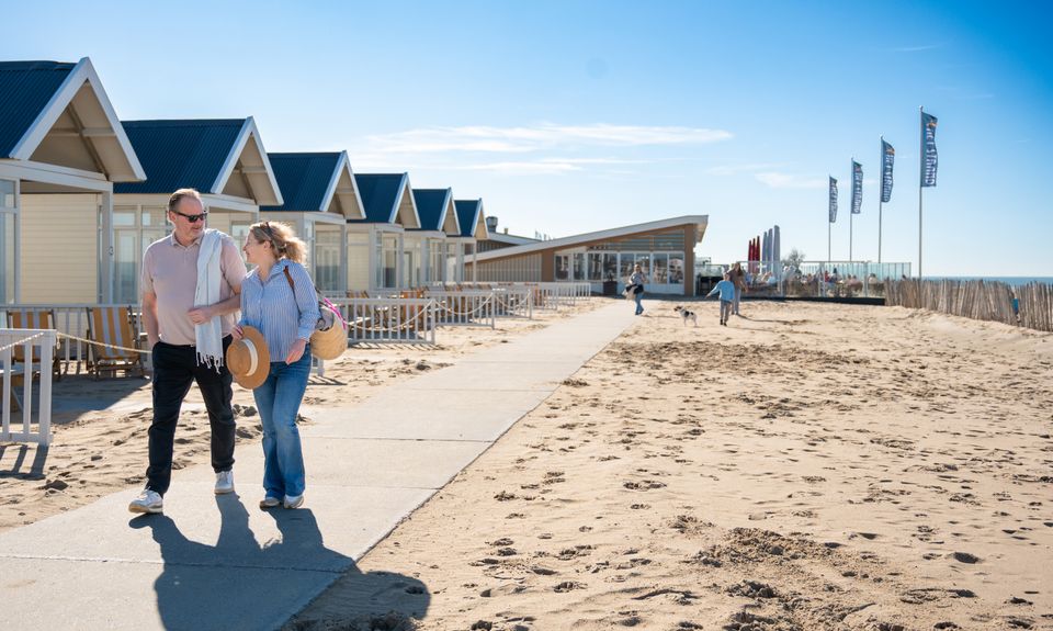 Een koppel loopt over het strand van Katwijk aan Zee, met op de achtergrond strandhuisjes en strandpaviljoen 'Het Strand' in beeld.