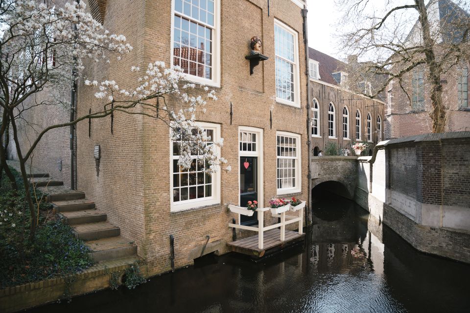Historisch museumcafé Museumtuin Gouda, Groene Hart Zuid-Holland, monumentaal bakstenen pand aan een gracht met klein terras aan het water, bloeiende magnoliaboom en brug naast de Sint-Janskerk in het centrum tijdens de lente.