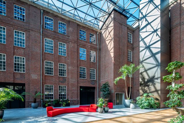 Foto van een met glas overdekte open ruimte (atrium). Zichtbaar zijn de ramen van de oude leerfabriek in de gevel, rode banken en groene planten in het midden.
