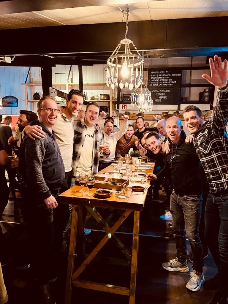 Groep volwassenen staat lachend en proostend rond een hoge tafel in een druk café. Er staan drankjes en hapjes op tafel. Informele sfeer met opgestoken armen en gezellig samenzijn tijdens een borrel.