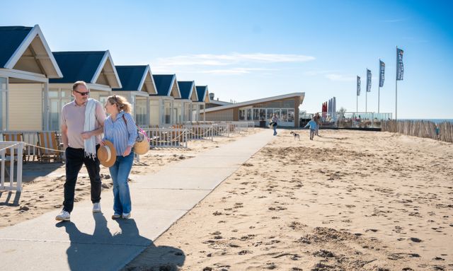 Een koppel loopt over het strand van Katwijk aan Zee, met op de achtergrond strandhuisjes en strandpaviljoen 'Het Strand' in beeld.
