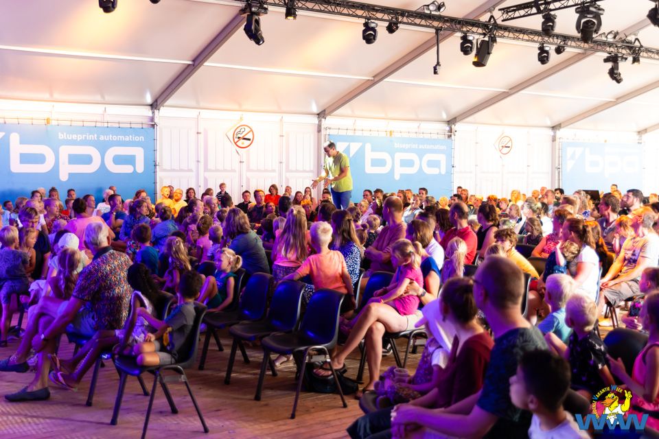 Een groep kinderen met ouders zittend op een stoeltje in een tent op de Woerdense VakantieWeek, kijkend naar kindertheater tijdens W-Celebrate.