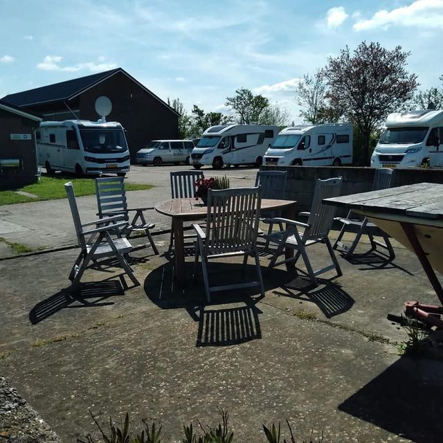 Terras met houten tafel en stoelen op Camperplaats Nieuw Vogelenzang in Montfoort, met op de achtergrond meerdere campers en een boerderijgebouw op een zonnige dag.