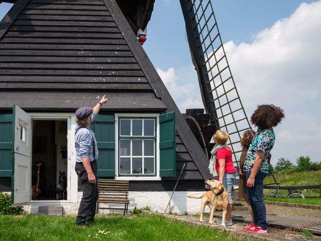 Drie personen staan bij een molen, de molenaar wijst omhoog.
