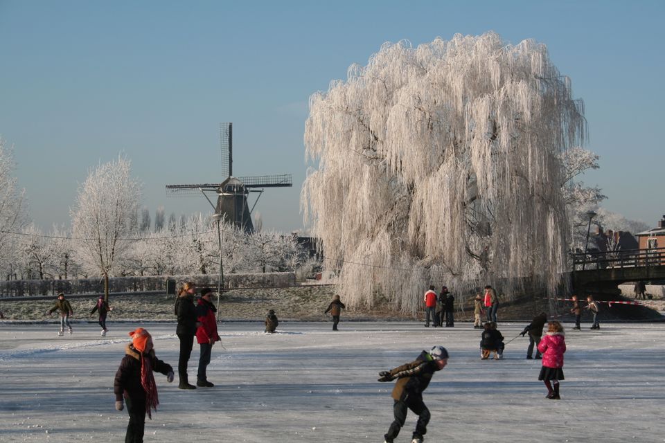 Twee kinderen maken het ijs speelklaar op een bevroren sloot: met bezems en een stoel improviseren ze hun eigen schaatsbaan. Op de achtergrond kijken buurtbewoners toe, terwijl het winterzonnetje de rijp op de bomen laat glinsteren. Een puur en herkenbaar winters moment waarin samen buiten zijn en creativiteit centraal staan.