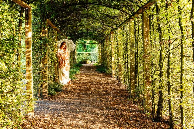 Vrouw staat met parasol onder begroeide overkapping waar het zonlicht doorheen filtert