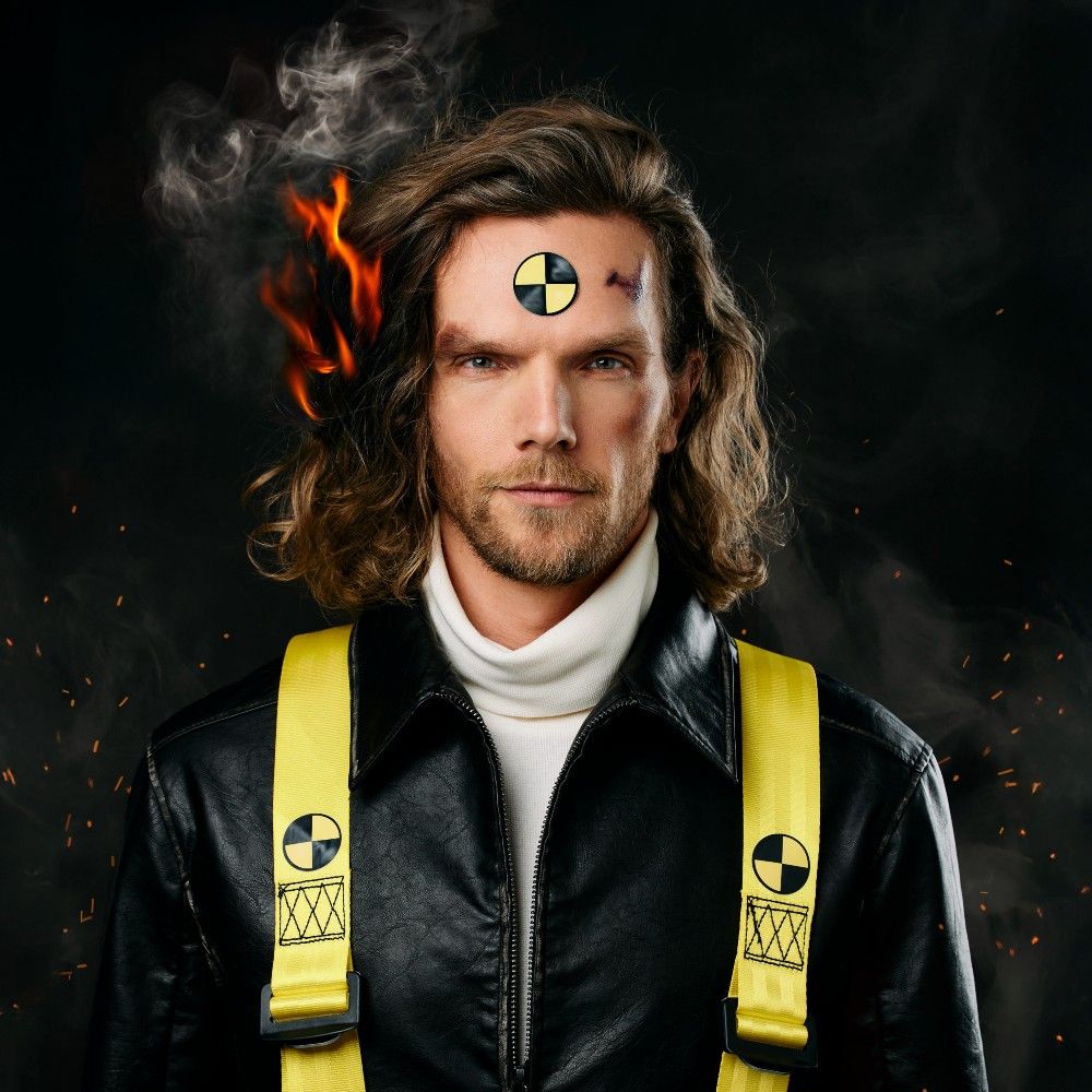 Gezicht van Ruud Smulders. Hij heeft een witte coltrui en een zwart jack met gele banden over de schouders aan. Langs zijn haren zijn vlammen te zien waardoor het lijkt of zijn haren in brand staat.