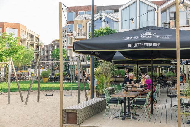 Gezellig terras bij het stadsstrand in Veenendaal, met zitplaatsen, groen en een kleine speeltuin. De foto laat zien hoe horeca en ontspanning samenkomen in een levendige stedelijke omgeving.
