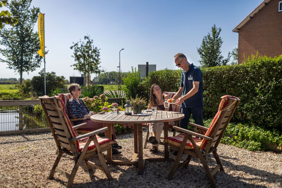 Bed & Breakfast Hazenveld in het Groene Hart, met gasten op het zonnige terras aan een houten tafel terwijl de gastheer eten serveert, omringd door groen en polderlandschap.