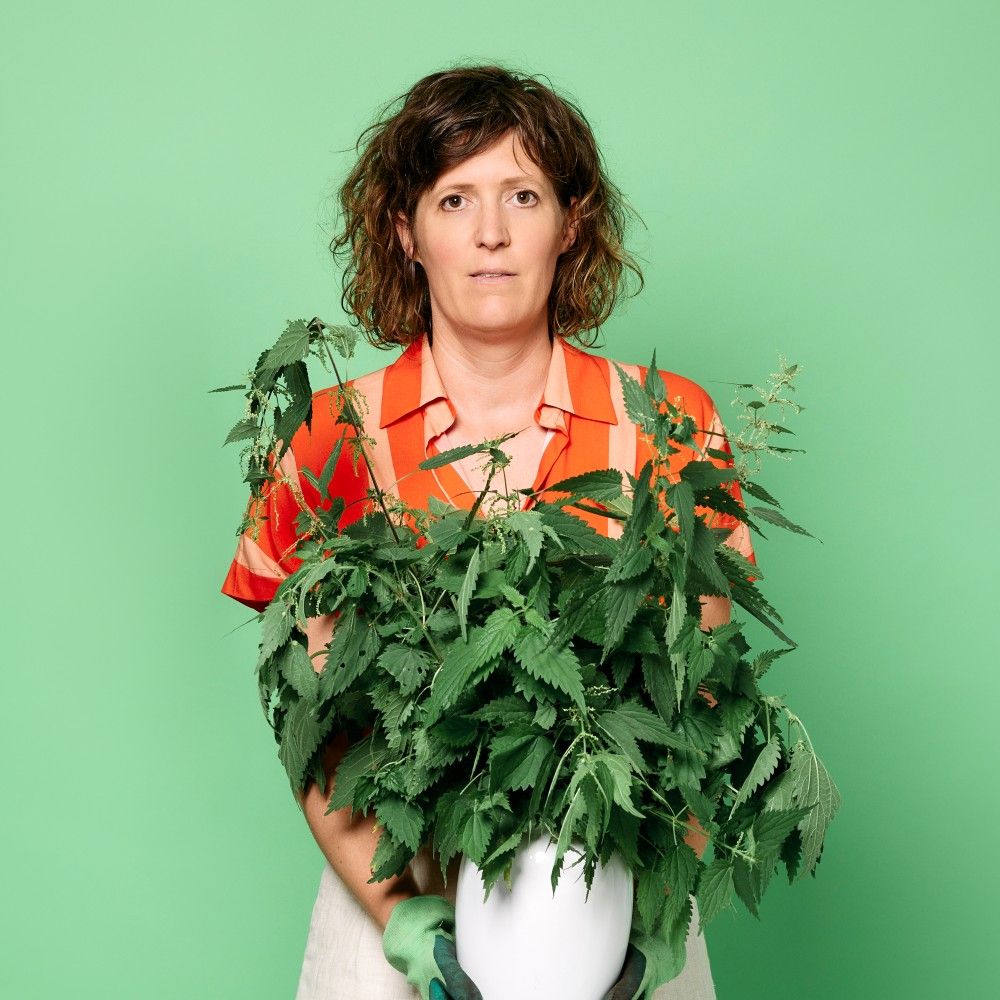 Foto van Janneke met een oranje bloesje aan tegen een groene achtergrond. Ze houdt een witte vaas met een grote bos brandnetels vast.