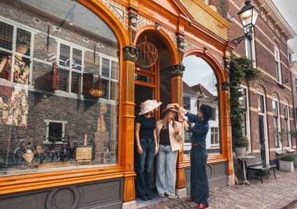 Hoedenatelier Beau Chapeau in Oudewater, Groene Hart. Ambachtelijke hoedenwinkel met historische gevel waar een hoedenmaakster twee bezoekers helpt met het passen van stijlvolle hoeden voor de entree in de sfeervolle binnenstad.
