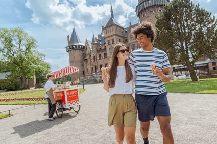 Twee bezoekers wandelen met een ijsje door de tuinen van Kasteel de Haar in Haarzuilens. Op de achtergrond staat het kasteel en een ijskar. Een zomerse dag in de historische kasteeltuinen van het Groene Hart.