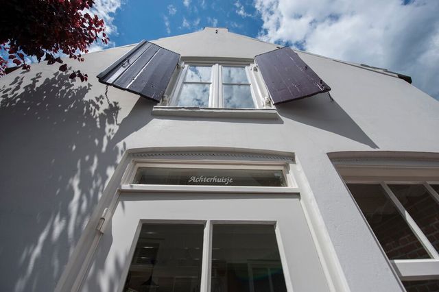 Witte gevelfoto van onderaf genomen met de voordeur en een raampje erboven met rode luiken. Boven de voordeur in het glas staat "Achterhuisje" gegraveerd.