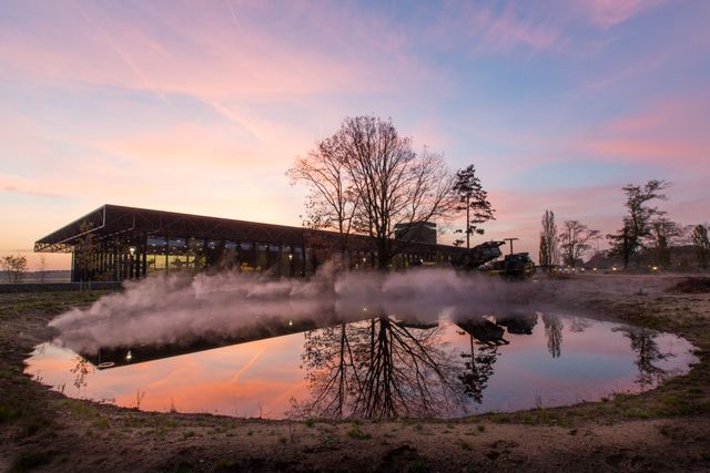 Het Nationaal Militair Museum in Soest bij zonsondergang, met roze avondlucht en weerspiegeling in het water. De sfeervolle foto toont het museumgebouw en militair materieel op het buitenterrein.